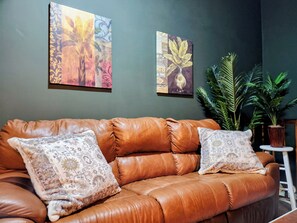 Ein braunes Ledersofa mit gemusterten Kissen, zwei an der Wand befestigte Kunstwerke und ein Topf mit Pflanze.