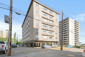 A multi-story building with a flat facade, several windows, and a parking area with cars. - The Golden Jewel • 1BR • Parking • AC • Mins to DT (Edmonton)