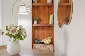 Une pièce avec un vase blanc contenant des fleurs, une chaise en osier, une bibliothèque en bois et un miroir.