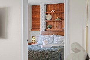 A bedroom with a wooden headboard, a bed with patterned bedding, a bedside lamp, and a clock on the wall. - The Golden Jewel • 1BR • Parking • AC • Mins to DT (Edmonton)