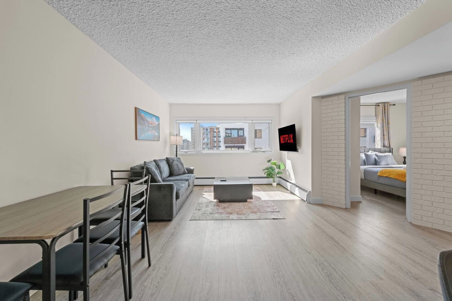 A modern living room with a sofa, coffee table, and TV. There is a dining area with a table and chairs. The room has a view of buildings outside the window.