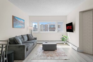 A modern living room with a grey sofa, a coffee table, a city view through the window, a Netflix sign, and a potted plant. - The Getaway • 2BR • Parking • AC • Mins to DT (Edmonton)