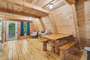 A wooden cabin interior with a dining table, benches, a sofa, and a window with curtains.