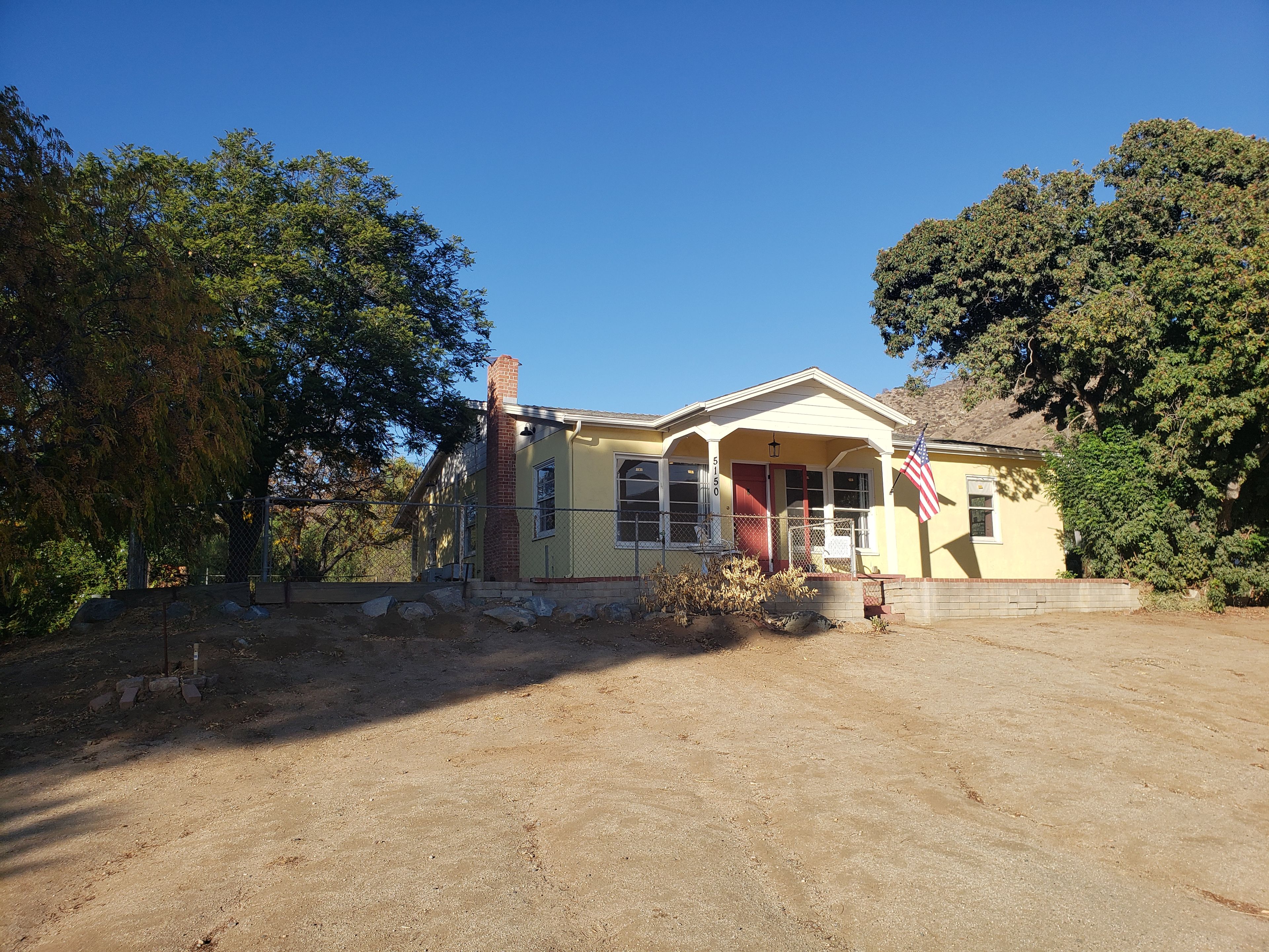 A single-story yellow house with a front porch, a brick chimney, and an American flag.