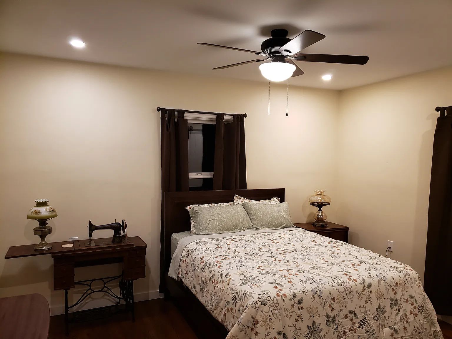 A bedroom with a bed, a desk, a sewing machine, a ceiling fan, and a nightstand with a lamp.