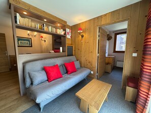 Ein gemütliches Zimmer mit einem grauen Sofa, roten Kissen, einem Holztisch und einem Bücherregal voller Bücher.