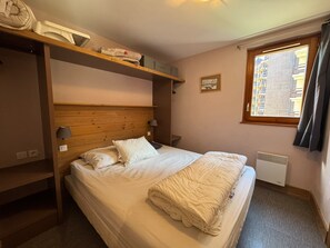 Ein Schlafzimmer mit einem Bett, einer Kopfstütze, einem Fenster, einem Heizkörper und einem Regal mit Gegenständen.