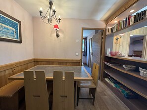 A dining area with a wooden table and benches, a chandelier, a bookshelf with books, and a framed picture on the wall. - Appartement T3 pour 7 personnes avec grand balcon - Résidence Antarès (Risoul)