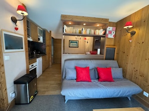 A compact room with a kitchenette, a sofa with red cushions, and a bookshelf filled with books. - Appartement T3 pour 7 personnes avec grand balcon - Résidence Antarès (Risoul)