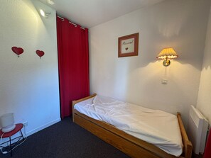 Quarto pequeno com uma cama de solteiro, estrutura de madeira, cortina vermelha, luminária na parede e um quadro.