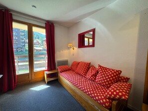Quarto com um banco de madeira, um sofá vermelho estampado, um espelho e uma janela com cortinas.