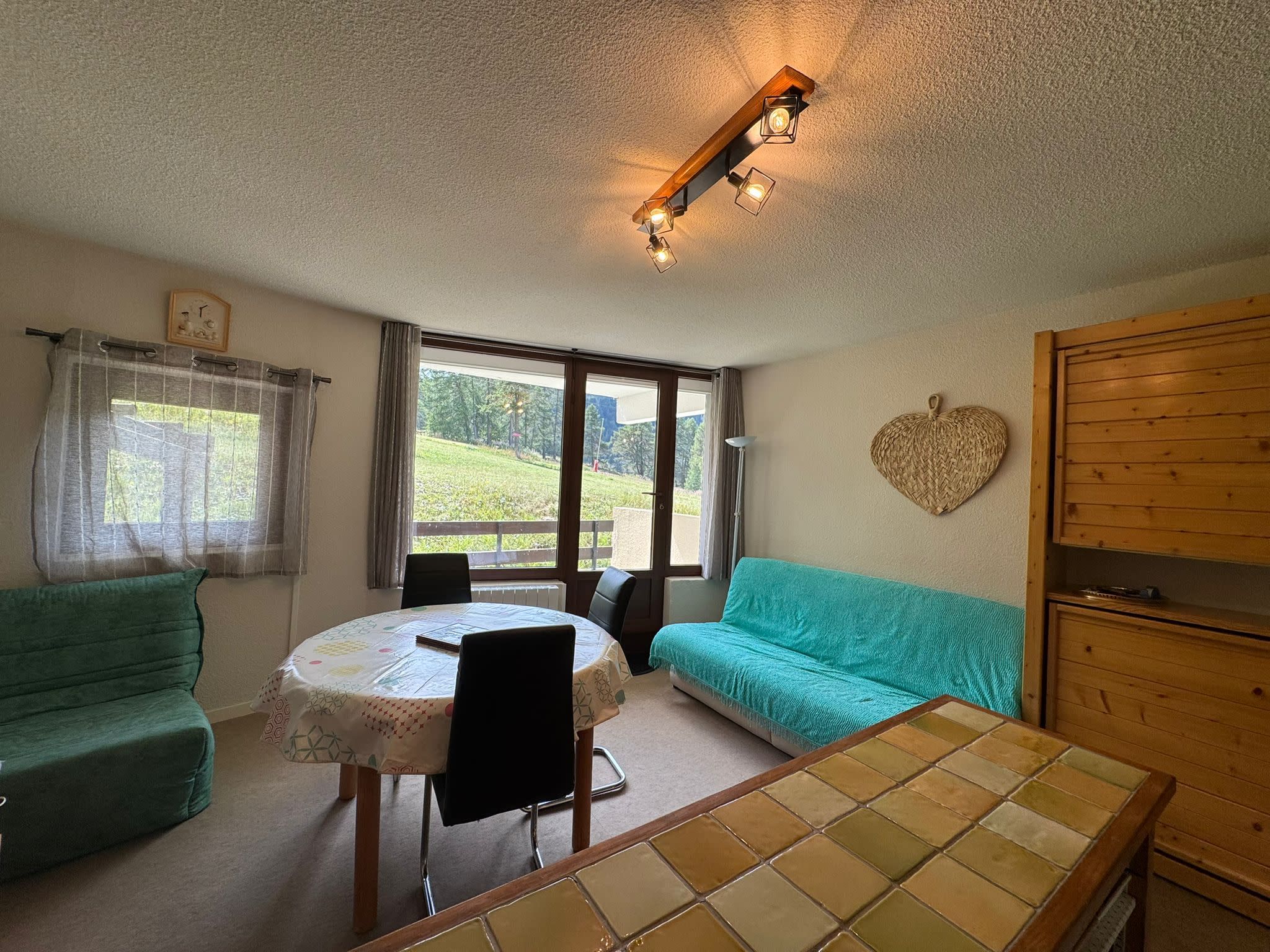 A cozy room with a round table, two green sofas, a wooden cabinet, and a large window offering a view of greenery.