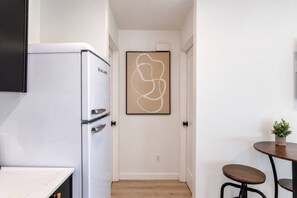 A compact kitchen with a white refrigerator, a door with a minimalist artwork, a small round table with a plant, and a wooden floor. - Cozy Casa | La Mesa Classic Craftsman (La Mesa)