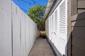 Ein schmaler Betonweg, gesäumt von einem weißen Holzzaun und einem Gebäude mit weißer Holzverkleidung und Fensterläden.