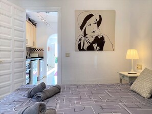 Ein Schlafzimmer mit einem Bett, einem Nachttisch, einer Lampe, einem Gemälde und Blick auf die Küche.