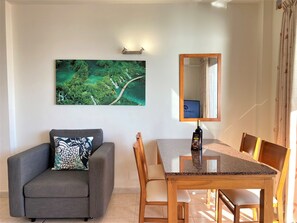 Ein Zimmer mit einem grauen Sofa, einem Holzesstisch mit Stühlen, einem großen Spiegel und einem Landschaftsbild mit grünen Bäumen.