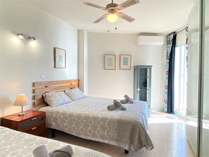 Ein Schlafzimmer mit Bett, Nachttisch, Lampe, Deckenventilator, Klimaanlage und Blick ins Freie.