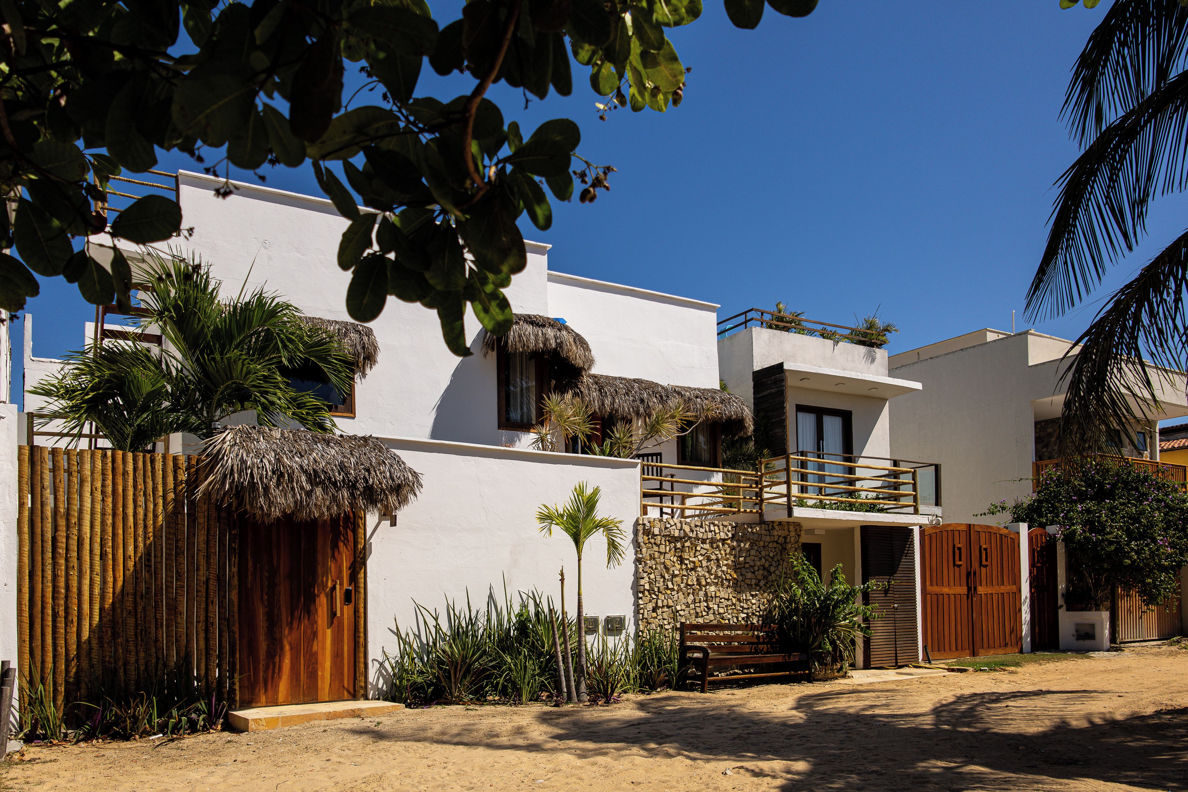 Una villa moderna con fachada blanca, techo de paja y balcón con vistas a una zona arenosa.