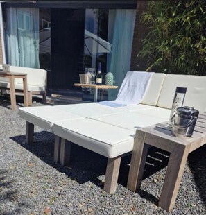 Une chaise longue avec un coussin, une petite table avec une bouteille et des verres, et un seau en métal posé sur une table en bois.