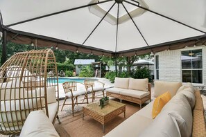 Un área de estar techada al aire libre con piscina, muebles de mimbre y un paraguas blanco.