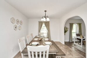 Un comedor con una mesa de madera, sillas blancas, una lámpara de araña y vistas a través de una ventana.