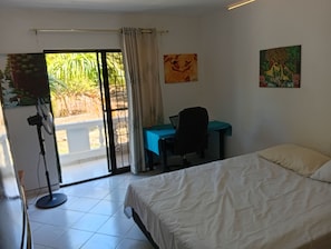 A bedroom with a bed, a desk, and a chair. There is a window with curtains, a fan, and a painting on the wall. - APARTMENT WITH A PRIVATE TERRACE (Sosúa)