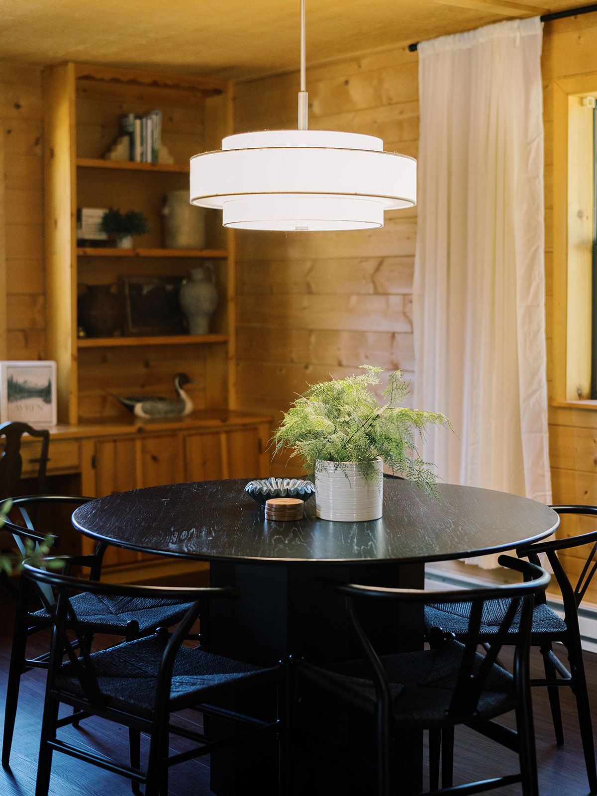 A round dining table with a potted plant, surrounded by chairs, in a room with wooden walls and a hanging light fixture.