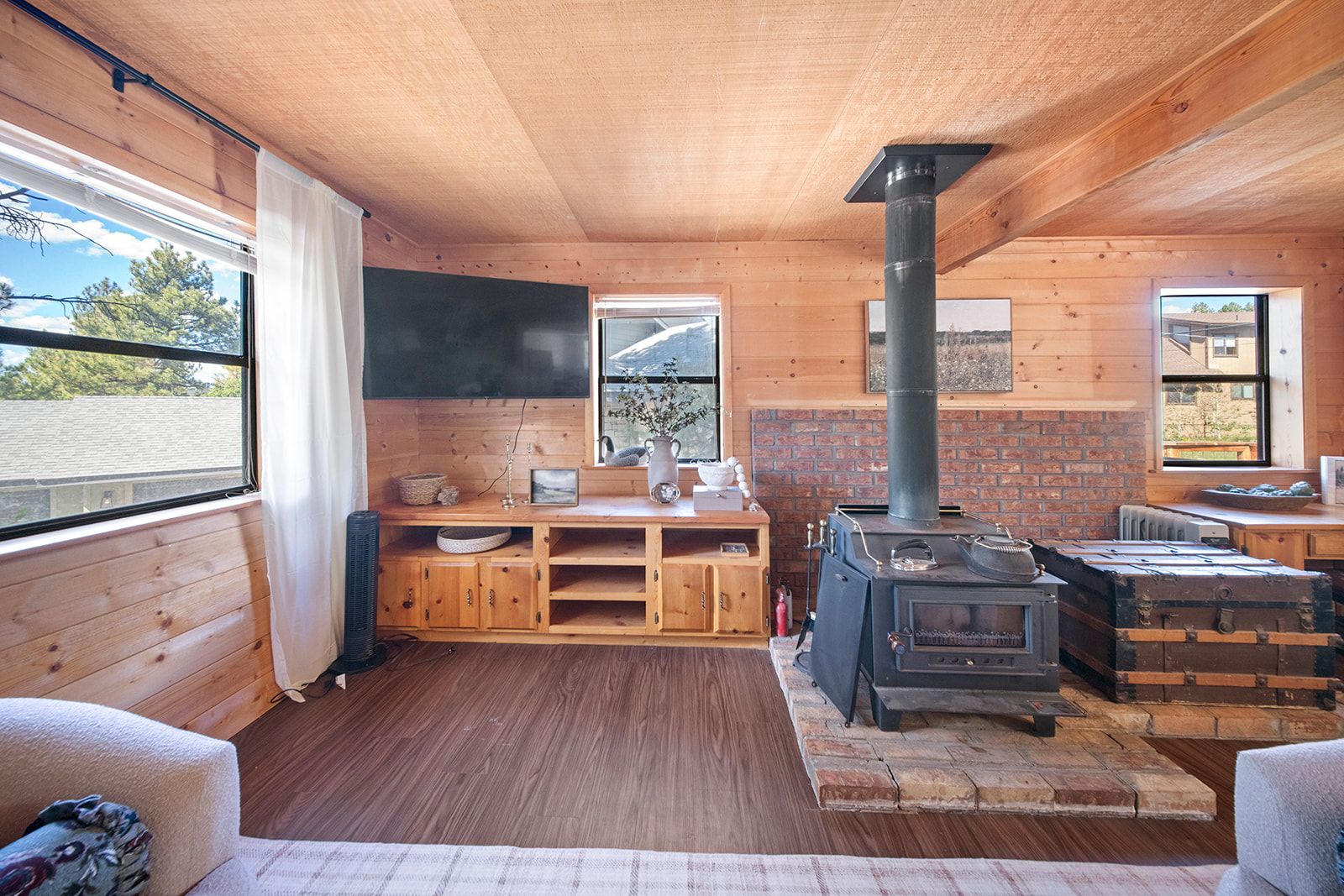 A cozy cabin interior with a wooden floor, a brick fireplace, and a large window providing a view of the outdoors.