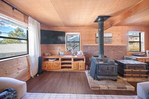 A cozy cabin interior with a wooden floor, a brick fireplace, and a large window providing a view of the outdoors.