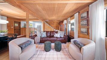 A cozy cabin interior with a wooden ceiling, a brown leather sofa, two armchairs, a dining table, and a kitchen area visible through an open door.