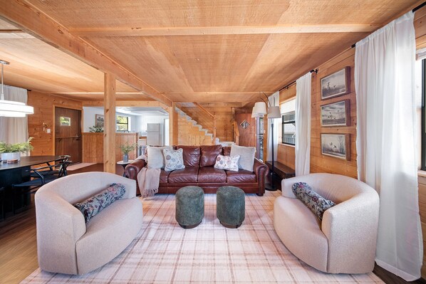 A cozy cabin interior with a wooden ceiling, a brown leather sofa, two armchairs, a dining table, and a kitchen area visible through an open door.