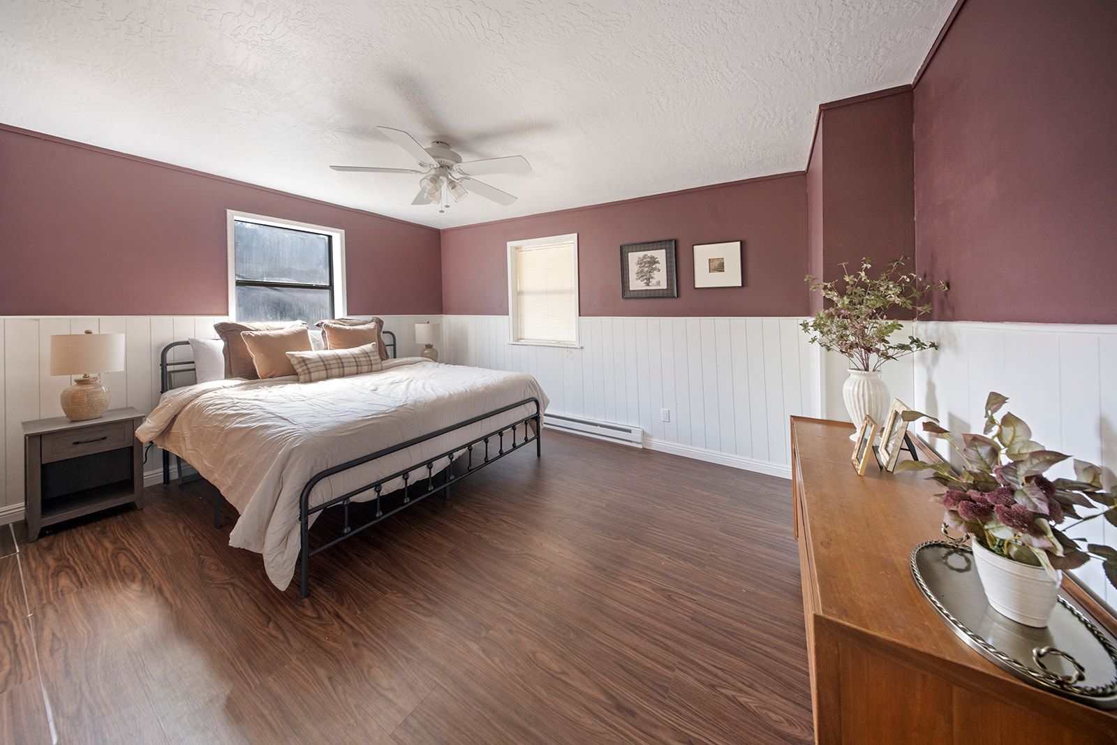 A bedroom with a bed, nightstand, lamp, ceiling fan, window, and wall-mounted pictures.