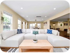 A spacious living room with a large L-shaped sectional sofa, a wooden coffee table, and a view of the adjacent dining area. - Houston Southwest Oasis – Heater Pool & Game Room (Houston)