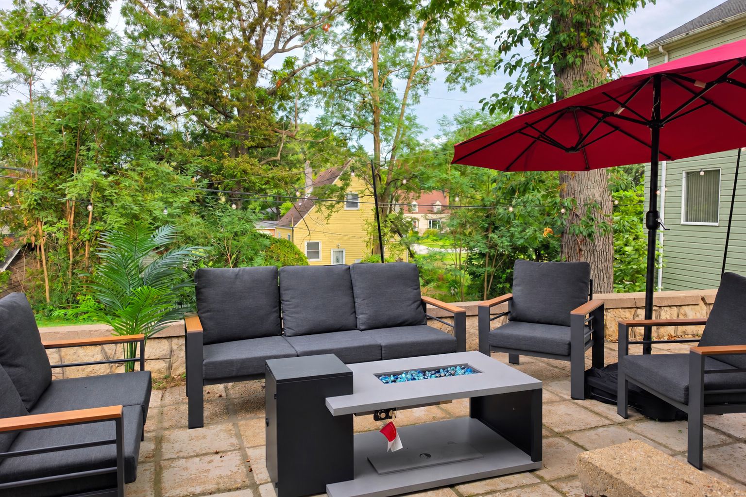 An outdoor seating area with a sofa, chairs, and a table, surrounded by greenery and a house in the background.