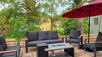 An outdoor seating area with a sofa, chairs, and a table, surrounded by greenery and a house in the background.