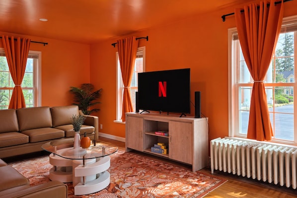 A living room with a sofa, a glass coffee table, a flat-screen TV, and orange curtains.