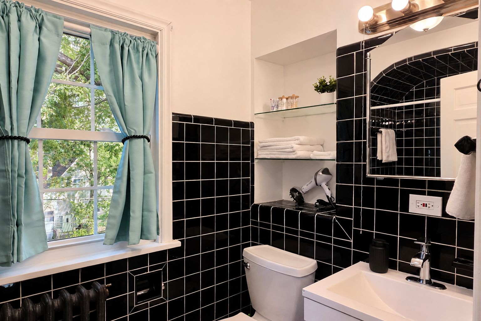 A bathroom with black tiled walls, a white toilet, a sink with a mirror, and a window with teal curtains.