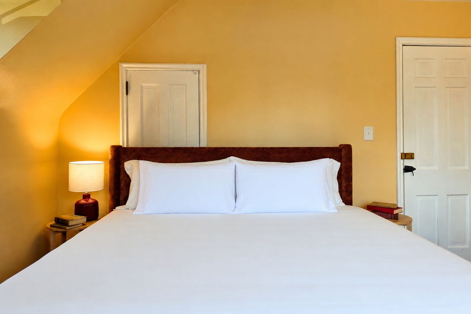A neatly made bed with white linens and a wooden headboard in a room with yellow walls and a sloped ceiling.
