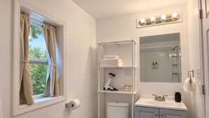 A bathroom with a toilet, sink, vanity, shelves with toiletries, a hairdryer, and a window with curtains. - Coastal Home | Patio & Firepit | Backyard Games (Merrillville)