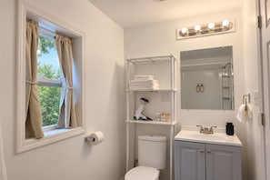 Ein Badezimmer mit Toilette, Waschbecken, Waschtisch, Regalen mit Toilettenartikeln, einem Föhn und einem Fenster mit Vorhängen.
