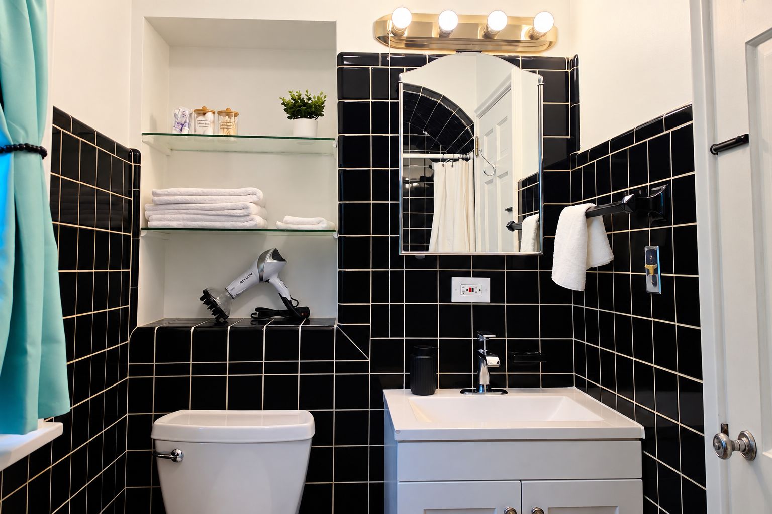 A bathroom with a toilet, sink, and a mirror with lights above it. There are towels on a shelf and a hairdryer on the counter.