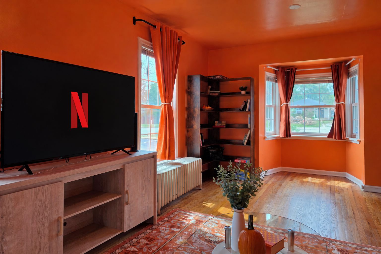 Een kamer met een grote flatscreen televisie, een boekenkast, een radiator en een raam met gordijnen.