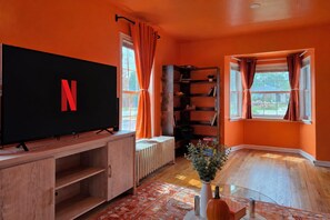 Ein Zimmer mit einem großen Flachbildfernseher, einem Bücherregal, einem Heizkörper und einem fenster mit Vorhängen.