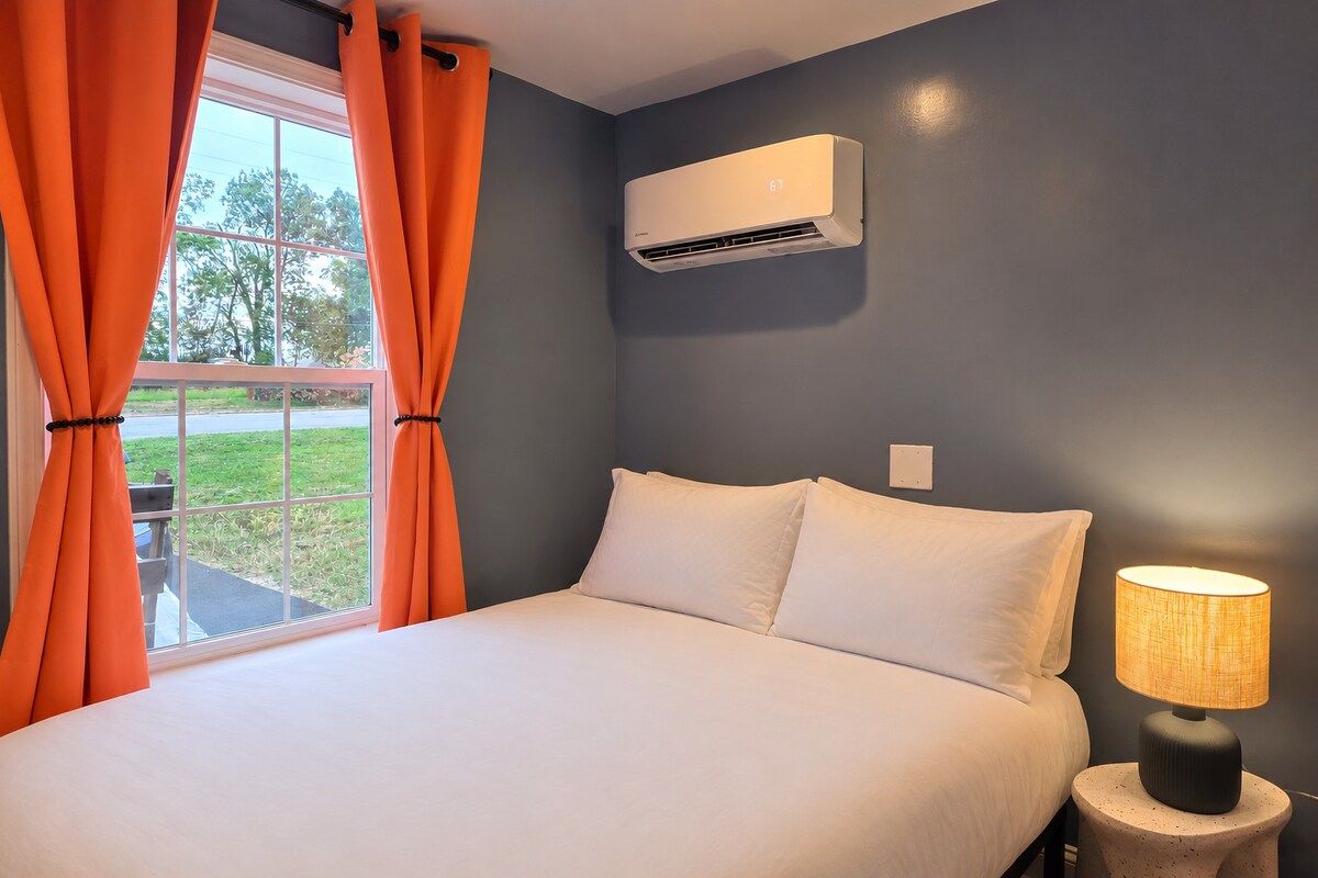 A bedroom with a bed, an air conditioning unit, a lamp, and orange curtains.