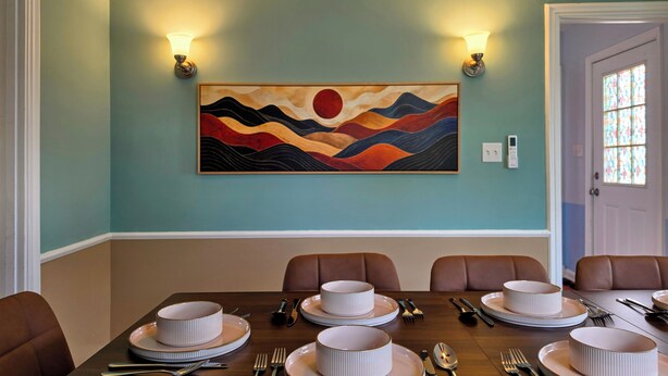 A dining area with a table set for four, a wall-mounted painting, and two wall sconces. - Coastal Home | Patio & Firepit | Backyard Games (Merrillville)