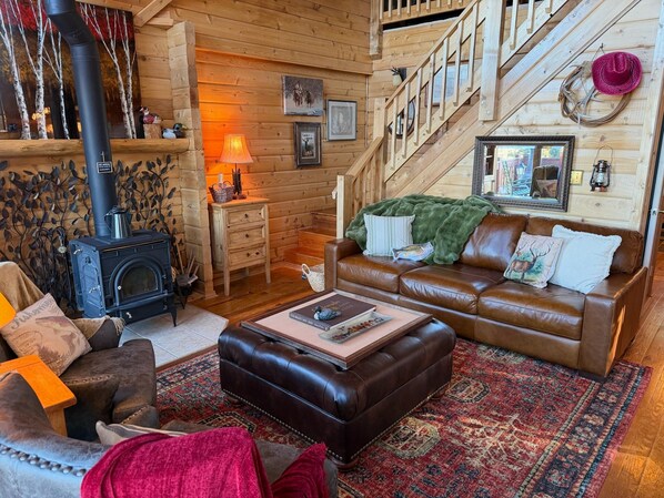 A cozy log cabin interior with a fireplace, leather furniture, and a staircase. - Cozy Cedar Cabin w/ Hot Tub, Mountain Views + Fireplace > Open Sky Cabin, CO (Jefferson)