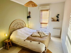 A bedroom with a bed, a bedside lamp, a window with blinds, a wall-mounted shelf with decorative items, and a standing heater. - A fully equipped haven of peace, close to downtown Aix-en-Provence, the TGV station, and the airport (Aix-en-Provence)