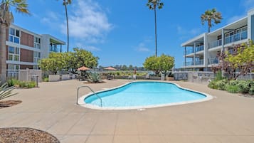 A swimming pool surrounded by buildings with balconies, palm trees, and outdoor seating.