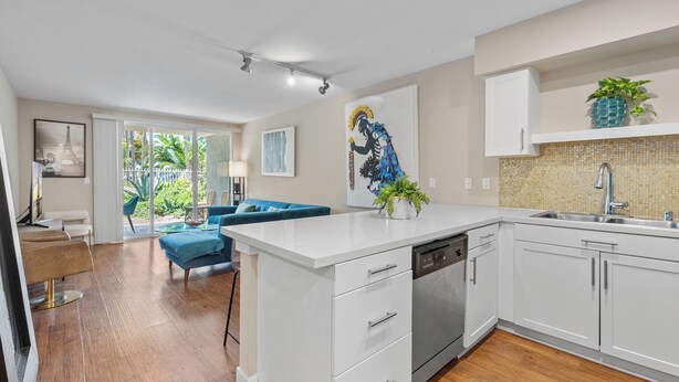 Una cocina moderna con muebles blancos, lavavajillas y un lavamanos. En la zona de estar contigua hay un sofá azul y una mesa de comedor.