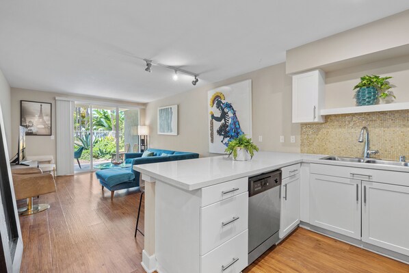 A modern kitchen with white cabinetry, a dishwasher, and a sink. There is a blue sofa and a dining table in the adjacent living area.
